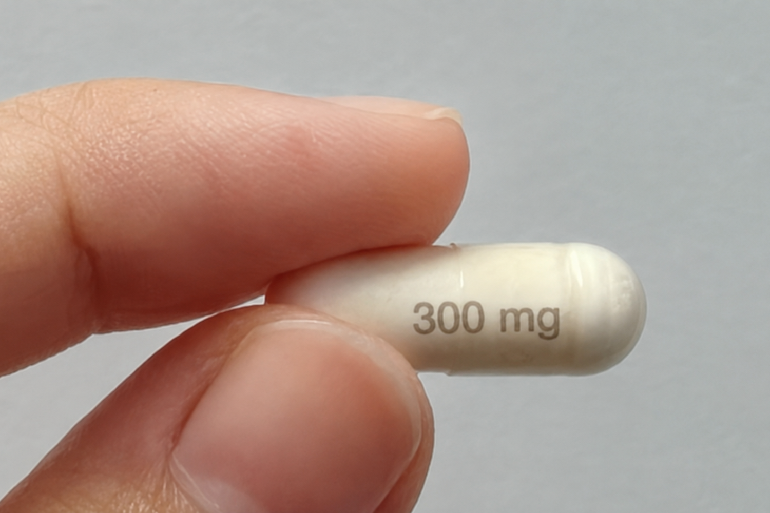 Close-up of a hand holding a small white capsule with “300 mg” printed on it against a light gray background.