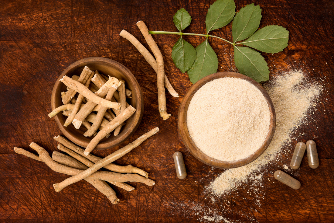 Ashwagandha root powder in a wooden bowl with dried roots, green leaves, and capsules on a wooden table.