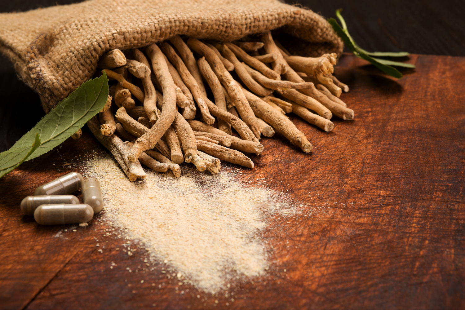 Ashwagandha roots spilling from a burlap sack with powder, green leaves, and capsules on a wooden table.