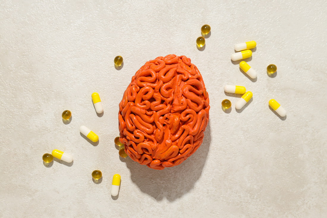 A model of a brain surrounded by yellow and white capsules and small yellow pills on a textured surface.