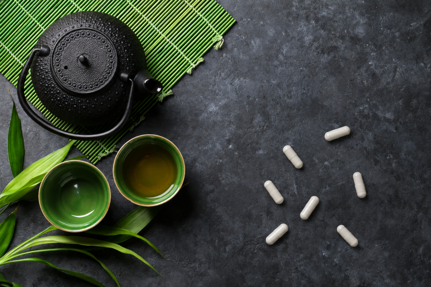 Top-down view of a black teapot, two green tea cups, and white supplement capsules on a dark surface.