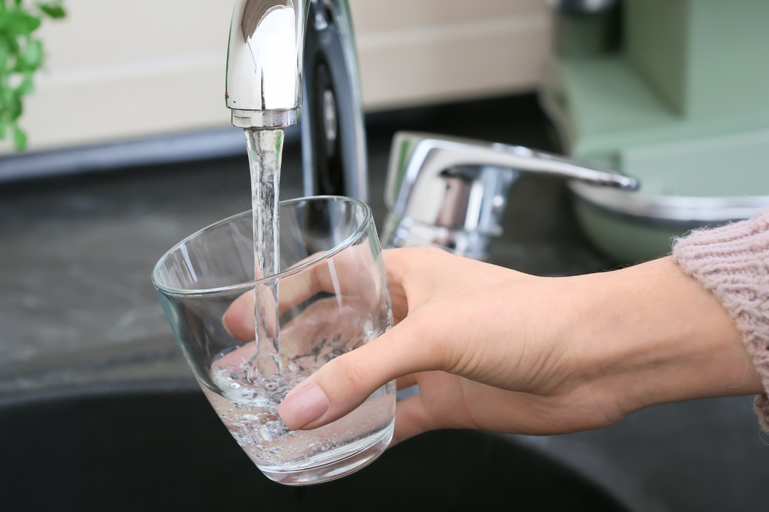 A hand holds a clear drinking glass under a chrome faucet as water pours into it in a modern kitchen sink area.