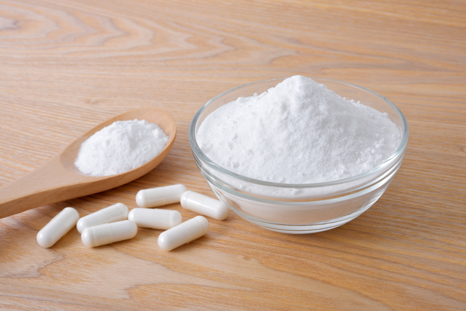 A small glass bowl and a wooden spoon filled with white lithium orotate powder sitting next to several white capsules on a light wooden table, representing the different supplement forms available for a beginner's dosage.