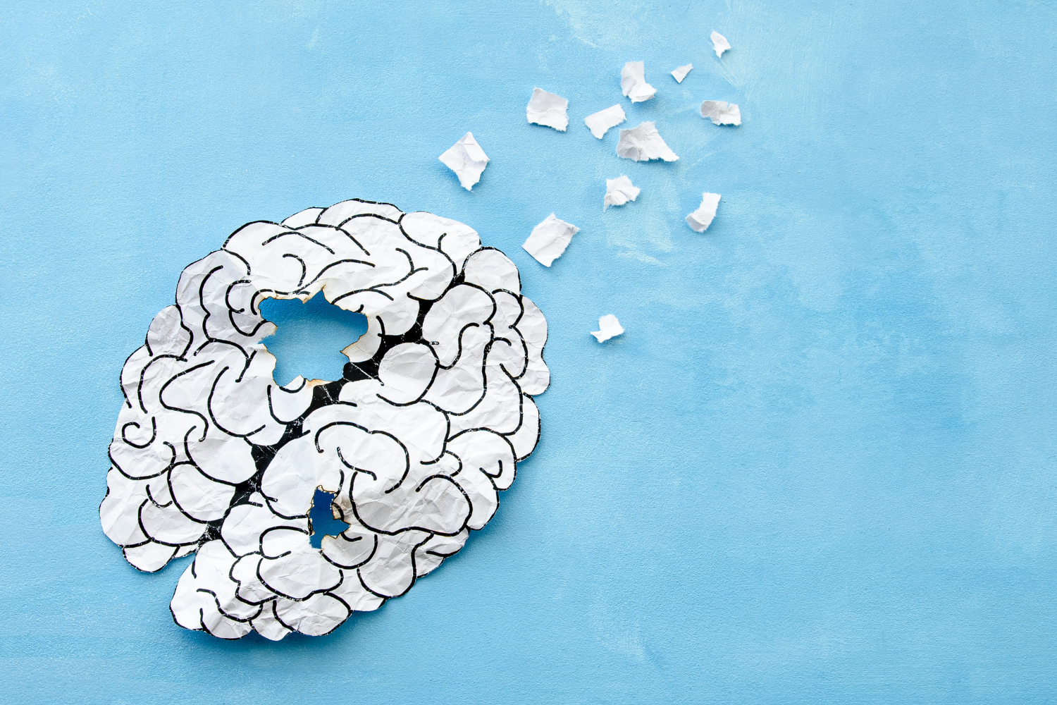 A paper cutout of a brain with pieces missing and scattered, on a blue background, signifying brain aging.