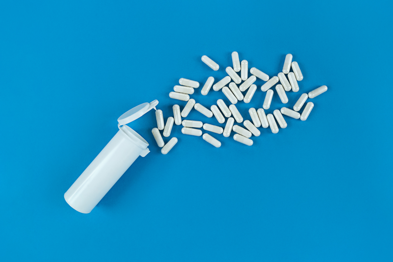 White dietary supplement capsules spilling from an open bottle on a blue background