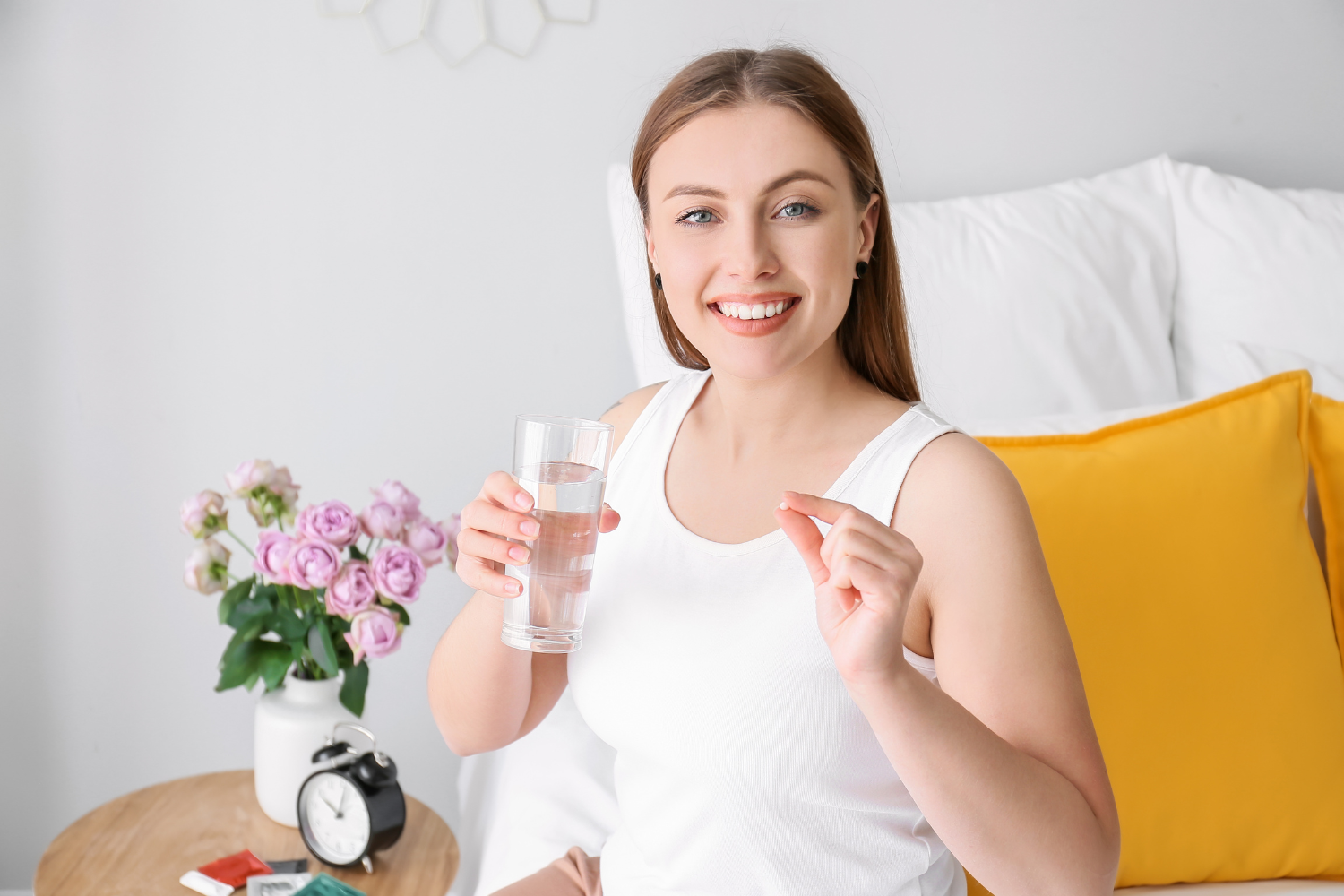 A smiling person holds a glass of water and a lithium orotate supplement, ready to take it. A vase of pink roses and an alarm clock are on a bedside table.